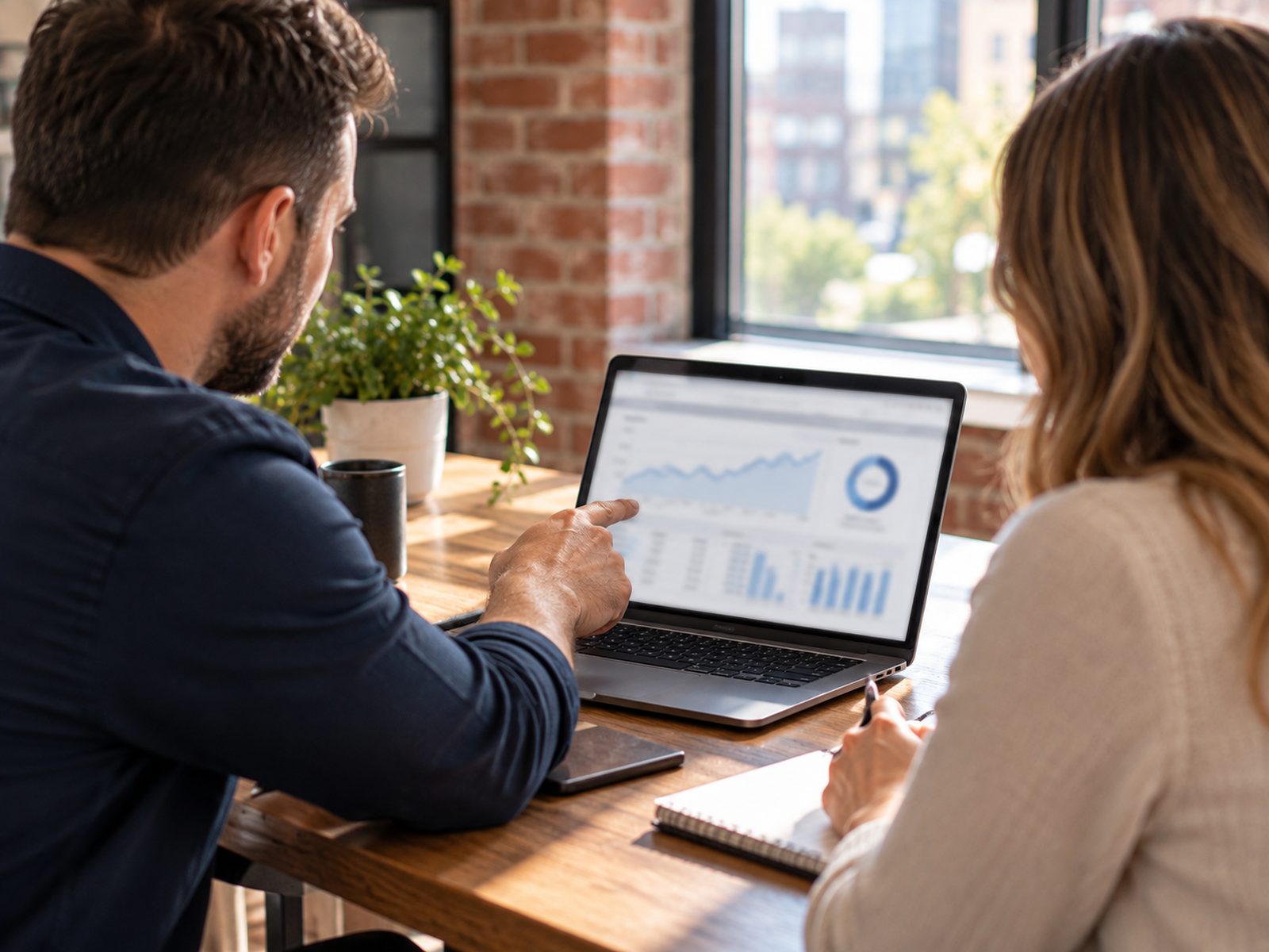 Two people reviewing a sales dashboard on a laptop in a brick-walled office